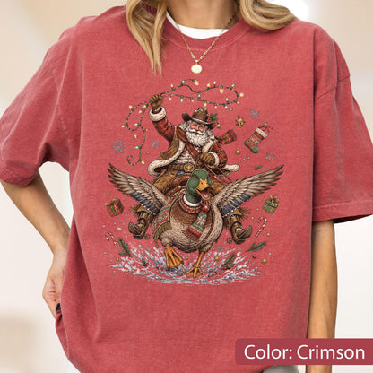 May include: A crimson short-sleeve t-shirt featuring a graphic print of Santa Claus wearing a cowboy hat and riding a flying mallard duck. The design includes festive elements like string lights, snowflakes, gift boxes, and candy canes.