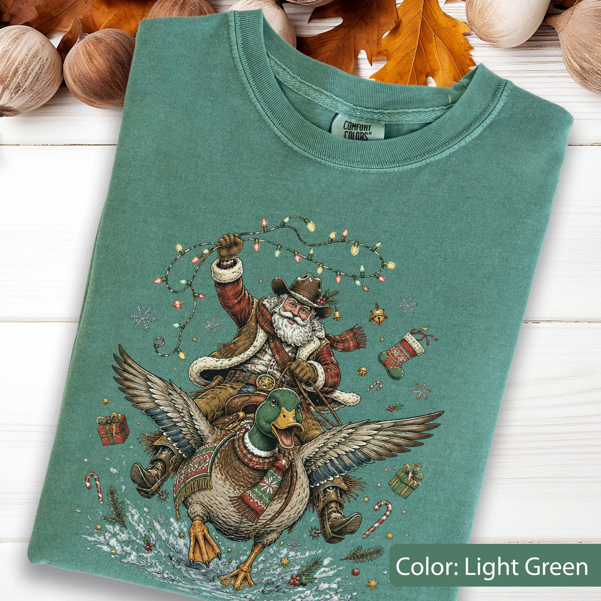 May include: A light green t-shirt featuring a graphic of a bearded figure in a cowboy hat and winter clothing riding a large mallard duck. The figure holds a string of colorful Christmas lights. The design includes small gifts, a stocking, and candy canes.