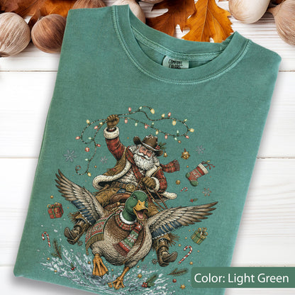 May include: A light green t-shirt featuring a graphic of a bearded figure in a cowboy hat and winter clothing riding a large mallard duck. The figure holds a string of colorful Christmas lights. The design includes small gifts, a stocking, and candy canes.
