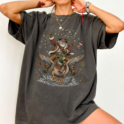May include: A charcoal gray short-sleeve t-shirt featuring a graphic print of Santa Claus wearing a cowboy hat and riding a flying mallard duck. The design includes Christmas lights, snowflakes, gift boxes, and a stocking surrounding the figures.