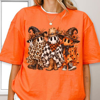 May include: An orange short-sleeve t-shirt featuring a graphic of three ghosts wearing hats and boots. The left ghost has a leopard print sheet, the middle ghost has a black and white checkered sheet, and the right ghost has an orange polka dot sheet.