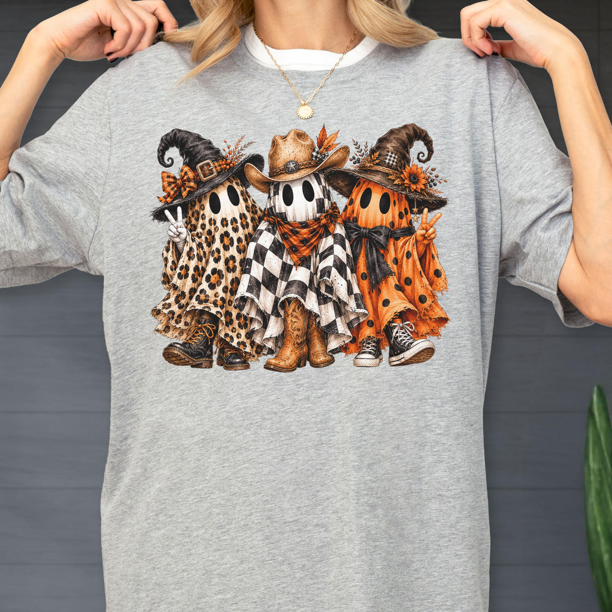 May include: A heather gray t-shirt featuring a graphic of three ghosts wearing hats and footwear. The left ghost has a leopard print sheet, the middle ghost has a black and white checkered sheet with a cowboy hat, and the right ghost has an orange polka dot sheet.