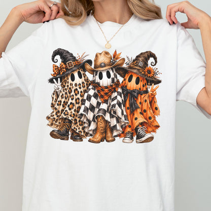May include: A white t-shirt featuring a graphic of three ghosts. The left ghost wears a leopard print sheet, a witch hat, and boots. The middle ghost wears a black and white checkered sheet, a cowboy hat, and boots. The right ghost wears an orange polka dot sheet, a witch hat, and sneakers.