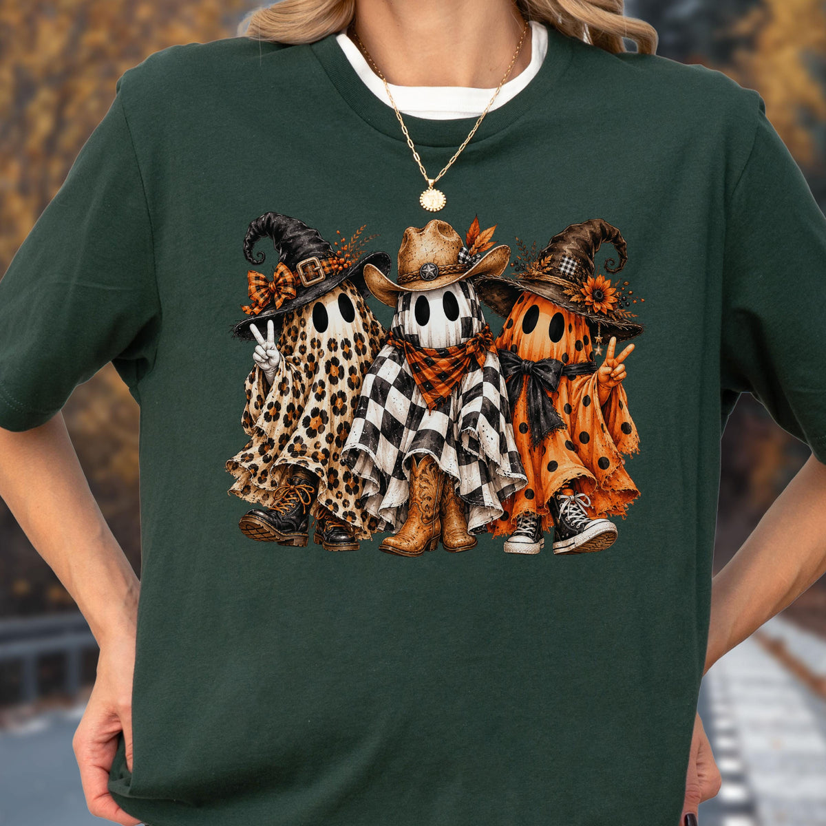 May include: A dark green short-sleeve t-shirt featuring a graphic of three ghosts wearing hats and footwear. The left ghost has a leopard print sheet, the middle ghost has a black and white checkered sheet, and the right ghost has an orange polka dot sheet.