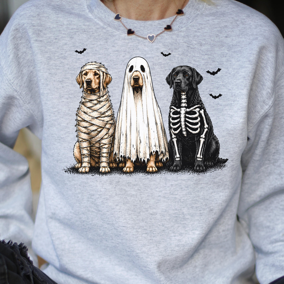 May include: A light gray crewneck sweatshirt featuring a graphic of three dogs in Halloween costumes: a golden retriever wrapped as a mummy, a golden retriever wearing a white ghost sheet, and a black labrador with a white skeleton print.