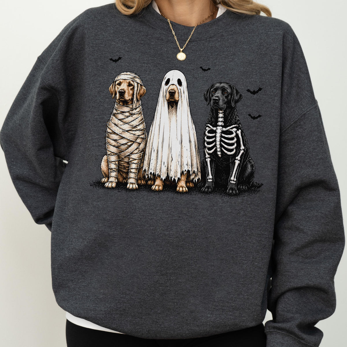 May include: A dark charcoal gray crewneck sweatshirt featuring a graphic print of three dogs in Halloween costumes: one wrapped like a mummy, one wearing a white ghost sheet, and one with a skeleton ribcage design, accented by small black bats.