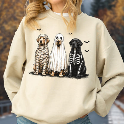 May include: A beige crewneck sweatshirt featuring a graphic of three dogs in Halloween costumes. One dog is wrapped like a mummy, one is wearing a white ghost sheet, and one has a white skeleton print on its black fur, with small black bats.