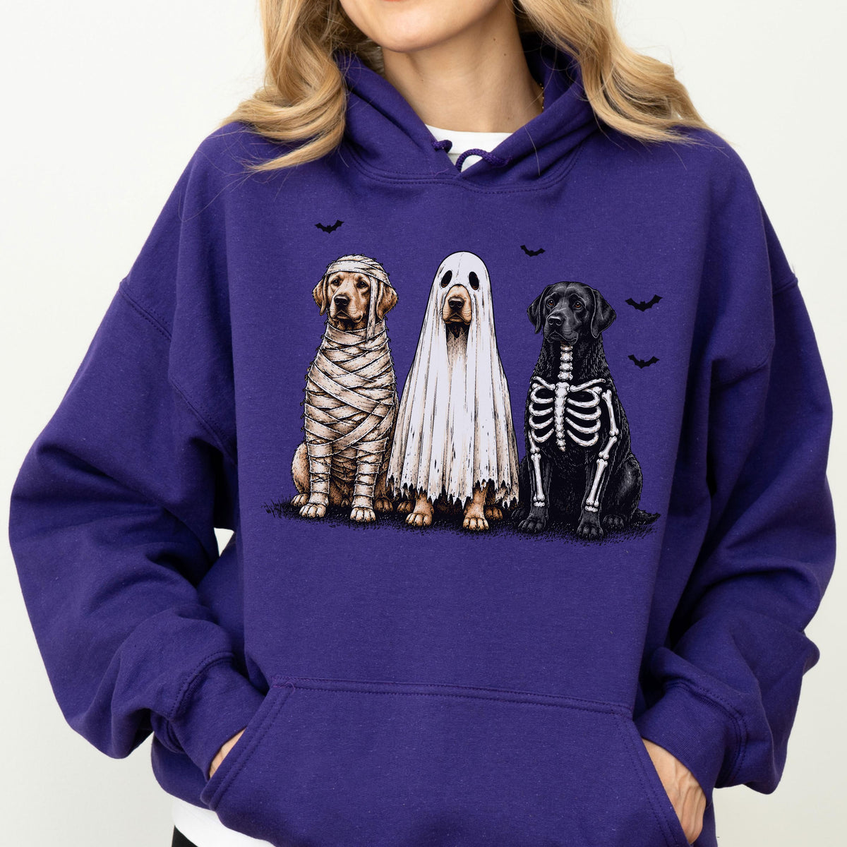 May include: A purple hooded sweatshirt featuring a graphic print of three dogs in Halloween costumes. One dog is wrapped like a mummy, one is wearing a white ghost sheet, and one has a skeleton bone pattern. Small black bats are printed above them.