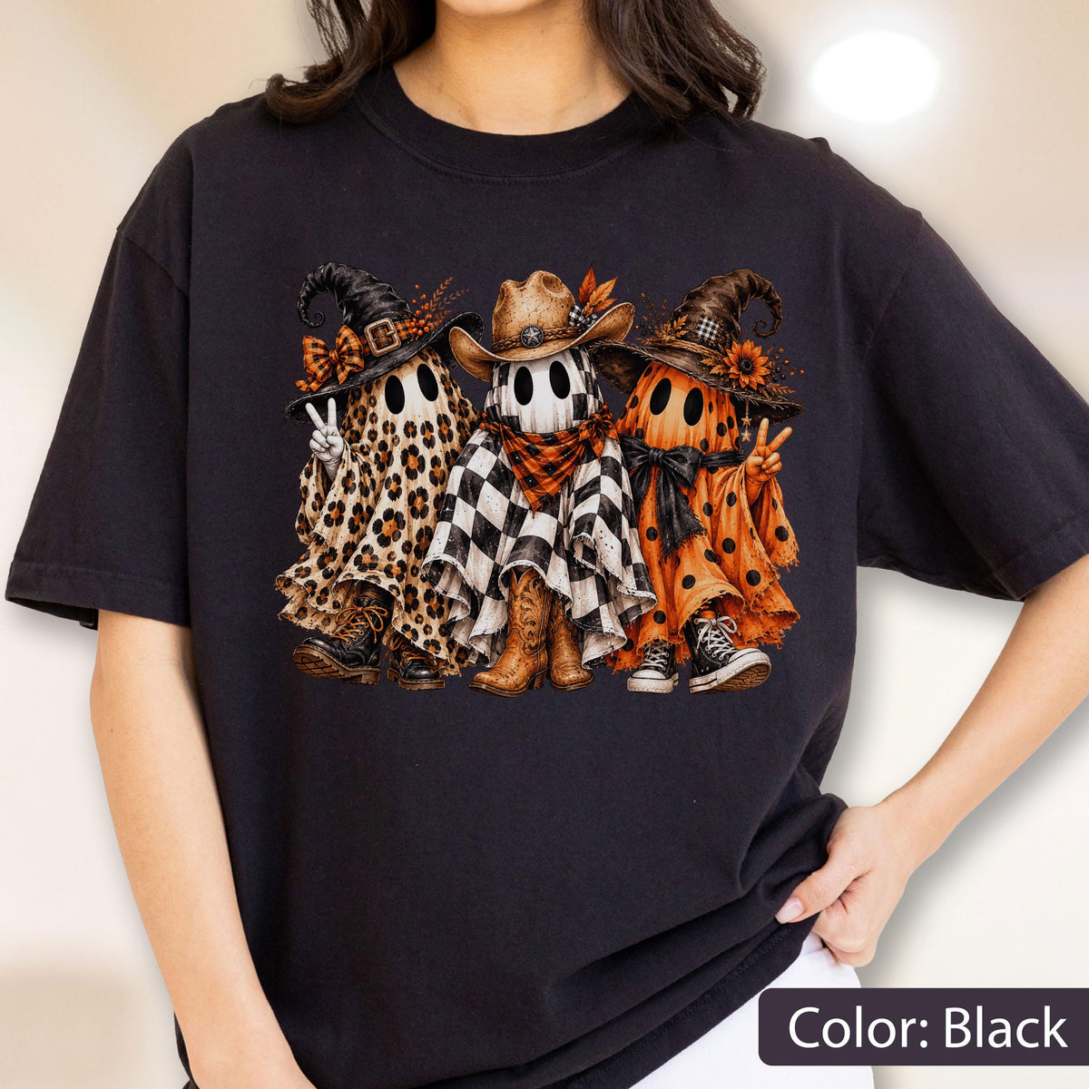 May include: A black t-shirt featuring a graphic of three ghosts wearing hats. The left ghost has a leopard print sheet, witch hat, and boots. The middle ghost wears a cowboy hat and checkered sheet. The right ghost has an orange polka dot sheet.