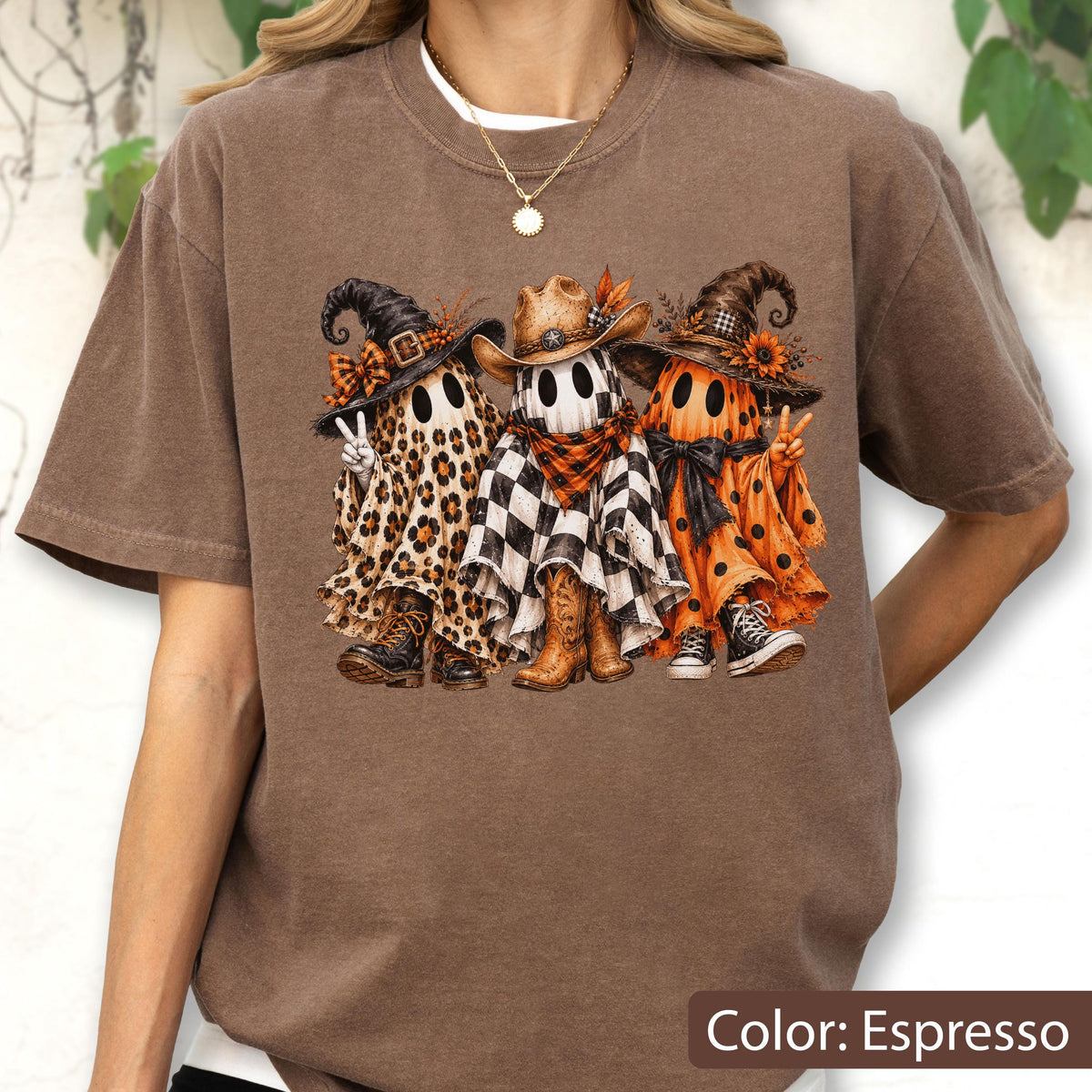 May include: An espresso-colored t-shirt featuring a graphic of three ghosts wearing hats. The left ghost has a leopard print sheet, the middle ghost has a black and white checkered sheet with a cowboy hat, and the right ghost has an orange polka dot sheet.