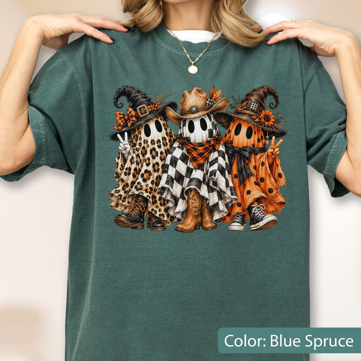 May include: A blue-spruce colored t-shirt featuring a graphic of three ghosts wearing hats. The left ghost has a leopard print sheet and combat boots, the center ghost has a checkered sheet and cowboy boots, and the right ghost has an orange polka dot sheet and sneakers.