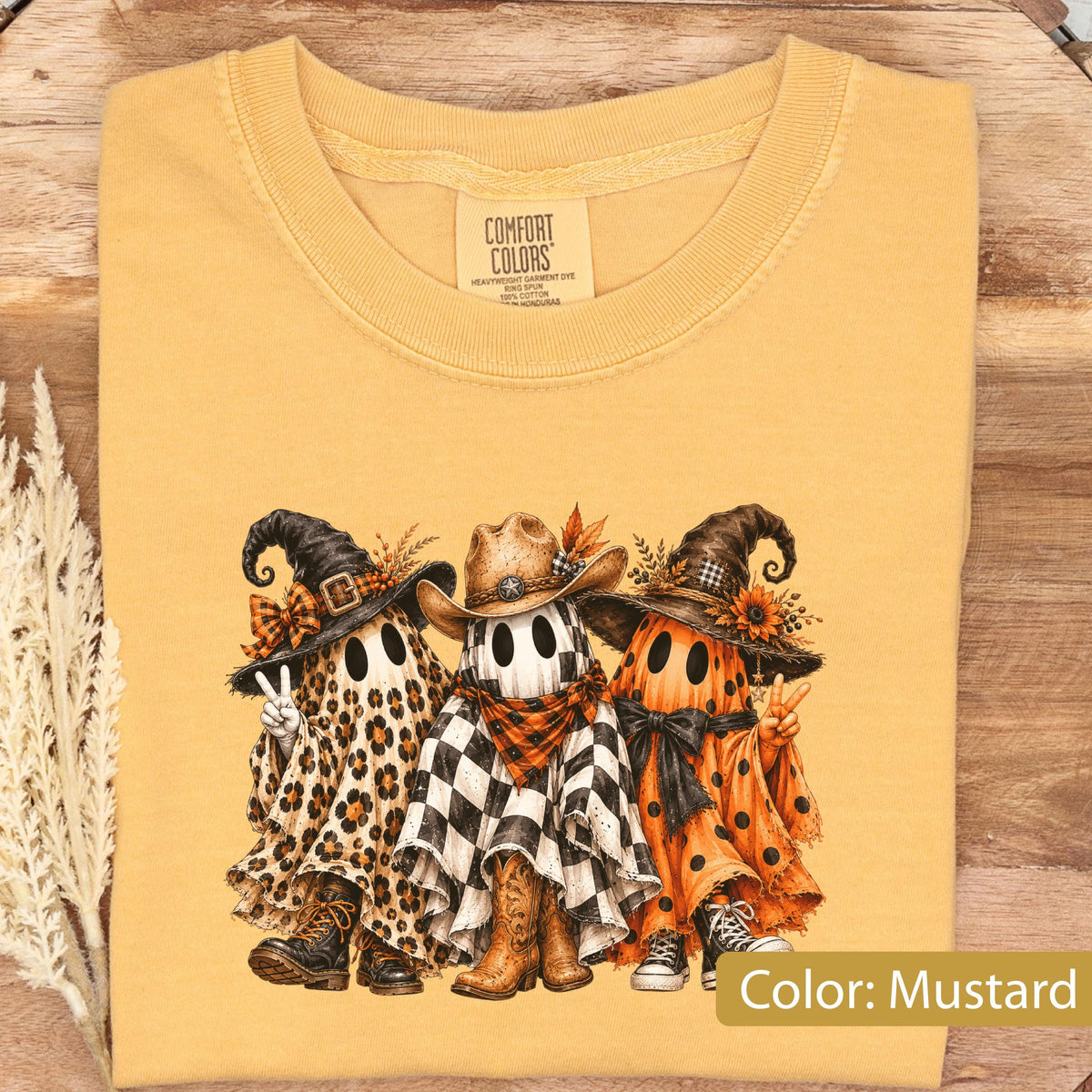May include: A mustard yellow t-shirt with a graphic print of three ghosts wearing hats. The left ghost wears a leopard print sheet and witch hat, the center ghost wears a checkered sheet and cowboy hat, and the right ghost wears a polka dot sheet.