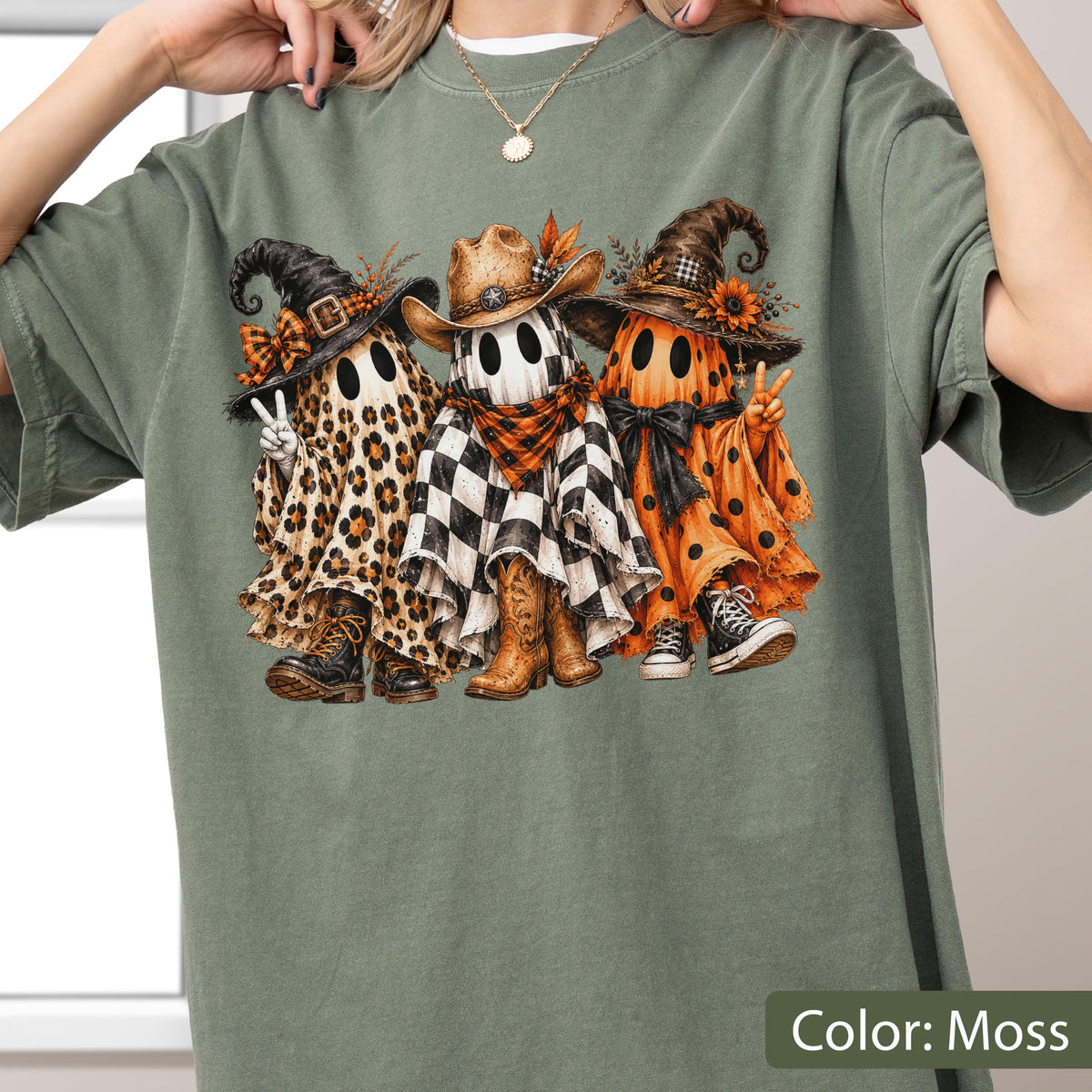 May include: A moss green t-shirt featuring a graphic of three ghosts wearing hats and footwear. The left ghost has a leopard print sheet, the middle ghost has a black and white checkered sheet with a cowboy hat, and the right ghost has an orange polka dot sheet.
