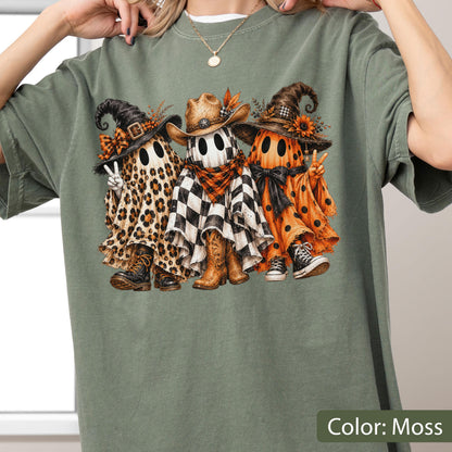 May include: A moss green t-shirt featuring a graphic of three ghosts wearing hats and footwear. The left ghost has a leopard print sheet, the middle ghost has a black and white checkered sheet with a cowboy hat, and the right ghost has an orange polka dot sheet.