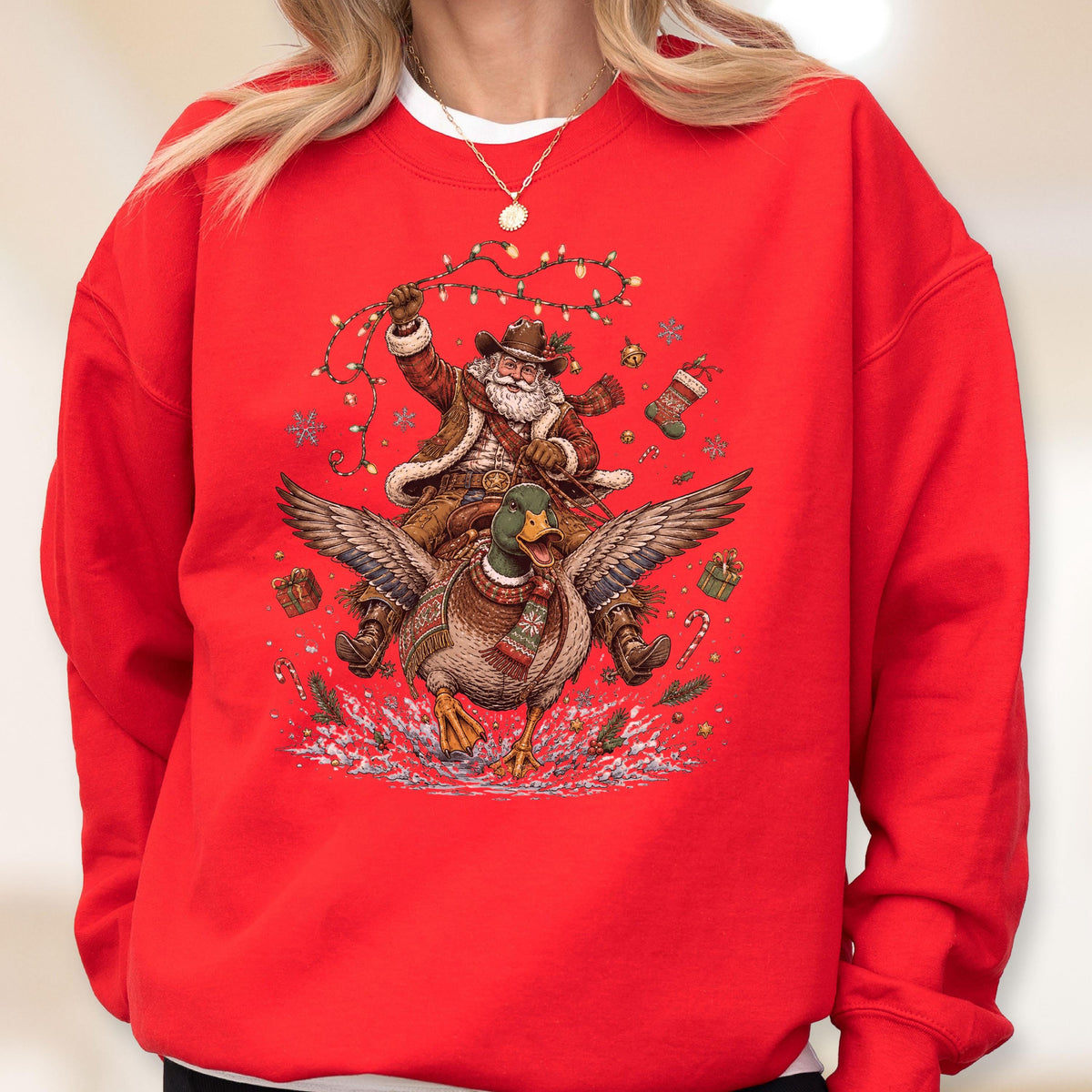 May include: A bright red crewneck sweatshirt featuring a graphic print of a cowboy Santa Claus riding a flying mallard duck. The design includes festive elements like string lights, a Christmas stocking, wrapped gifts, and candy canes.