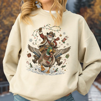May include: A beige crewneck sweatshirt featuring a graphic of Santa Claus wearing a cowboy hat and scarf while riding a flying mallard duck. The design includes Christmas lights, snowflakes, candy canes, and gift boxes surrounding the figures.