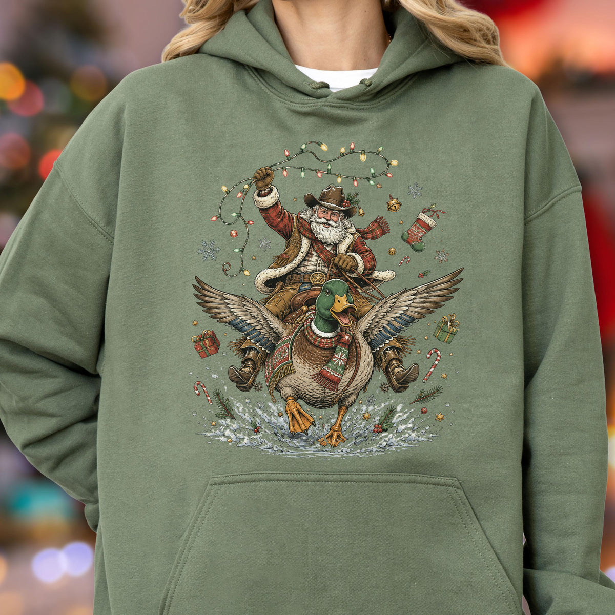 May include: An olive green hooded sweatshirt featuring a graphic print of Santa Claus wearing a cowboy hat and riding a mallard duck. Santa holds a string of colorful Christmas lights, surrounded by floating gift boxes, candy canes, and snowflakes.