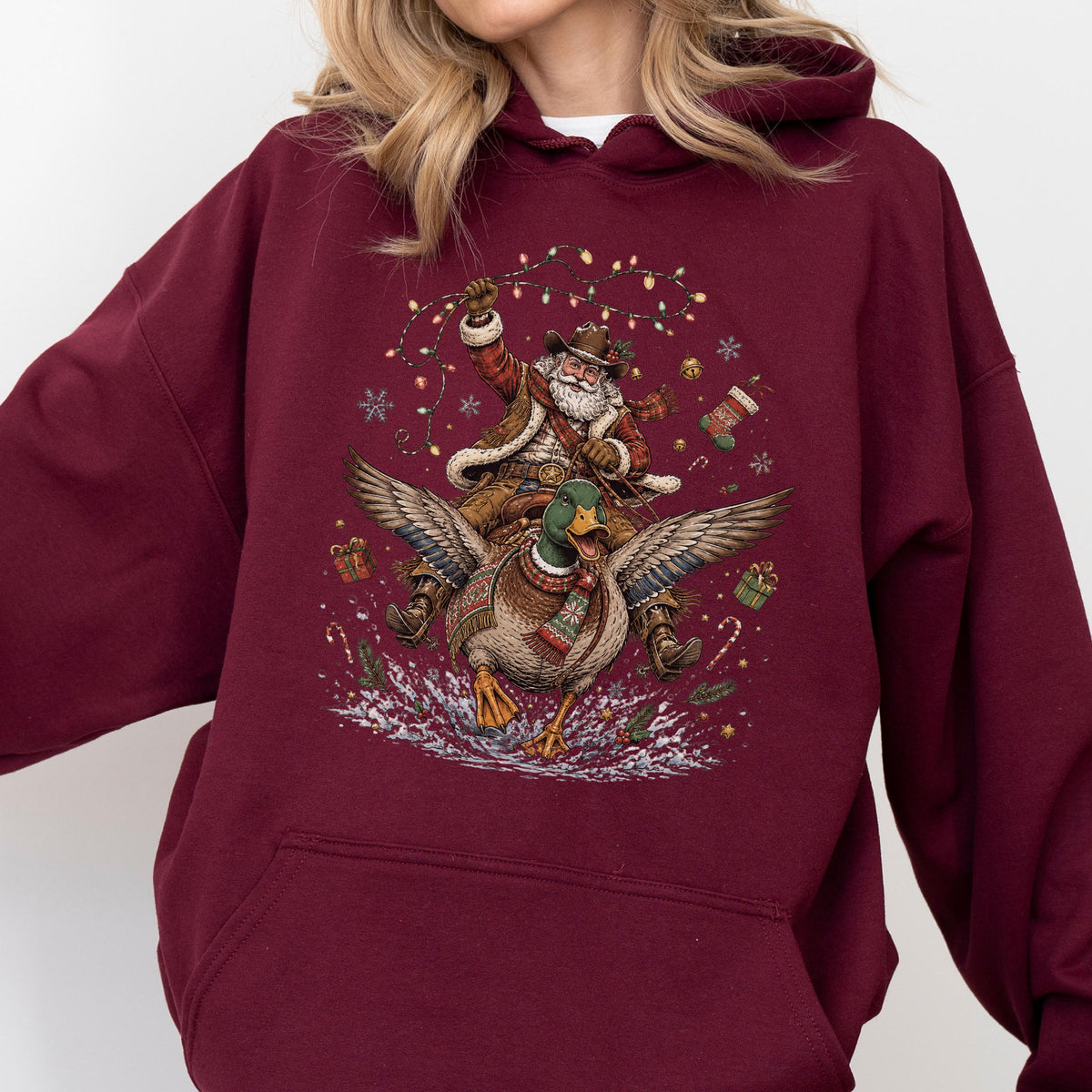 May include: A maroon hooded sweatshirt featuring a graphic print of Santa Claus riding a flying mallard duck. The design includes Christmas lights, snowflakes, wrapped gifts, and candy canes surrounding the central figures on the front of the garment.