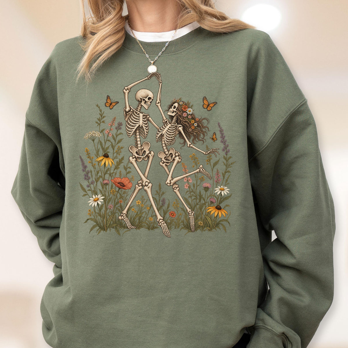 May include: An olive green crewneck sweatshirt featuring a graphic print of two skeletons dancing in a field of colorful wildflowers. The skeletons are surrounded by orange butterflies, daisies, poppies, and tall green grasses on the front chest.
