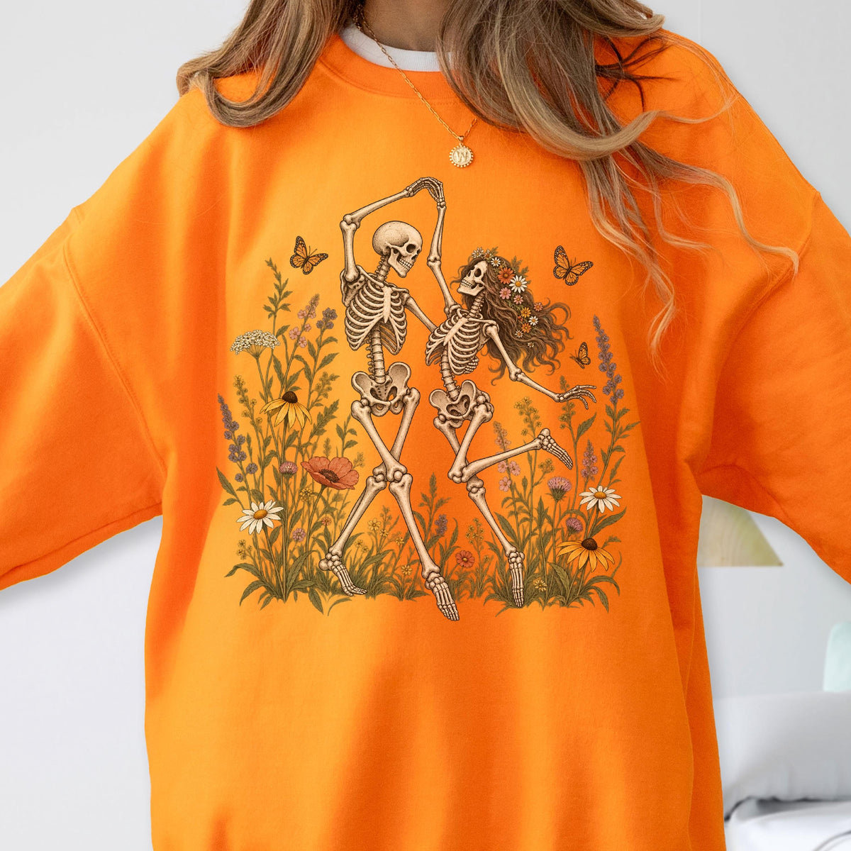 May include: A bright orange crewneck sweatshirt featuring a graphic print of two skeletons dancing in a field of wildflowers. The skeletons are surrounded by various colorful flowers, green foliage, and several butterflies in a detailed illustration.