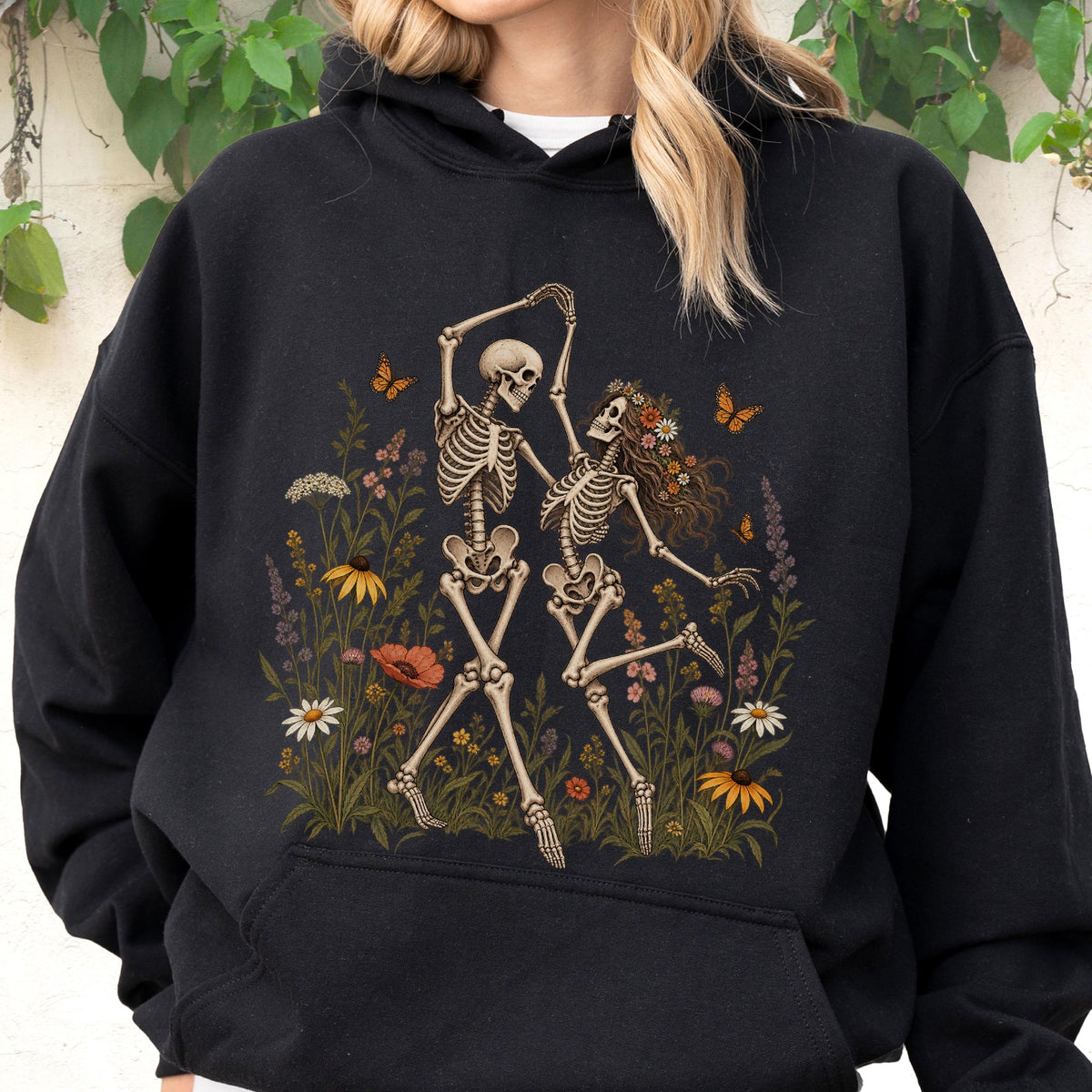 May include: A black hooded sweatshirt featuring a graphic print of two skeletons dancing in a field of colorful wildflowers. The skeletons are surrounded by various blossoms, green foliage, and orange butterflies, with one skeleton wearing a floral crown.