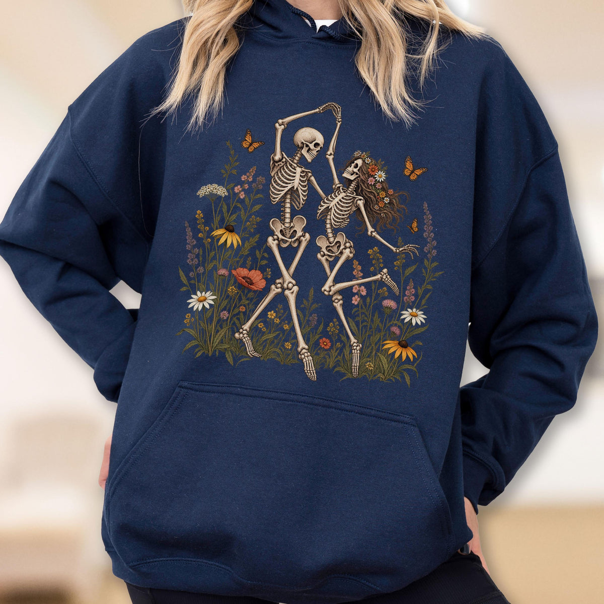 May include: A navy blue hooded sweatshirt featuring a graphic print of two skeletons dancing in a field of colorful wildflowers and butterflies. The design includes various floral elements in shades of yellow, red, white, and purple, with orange butterflies.