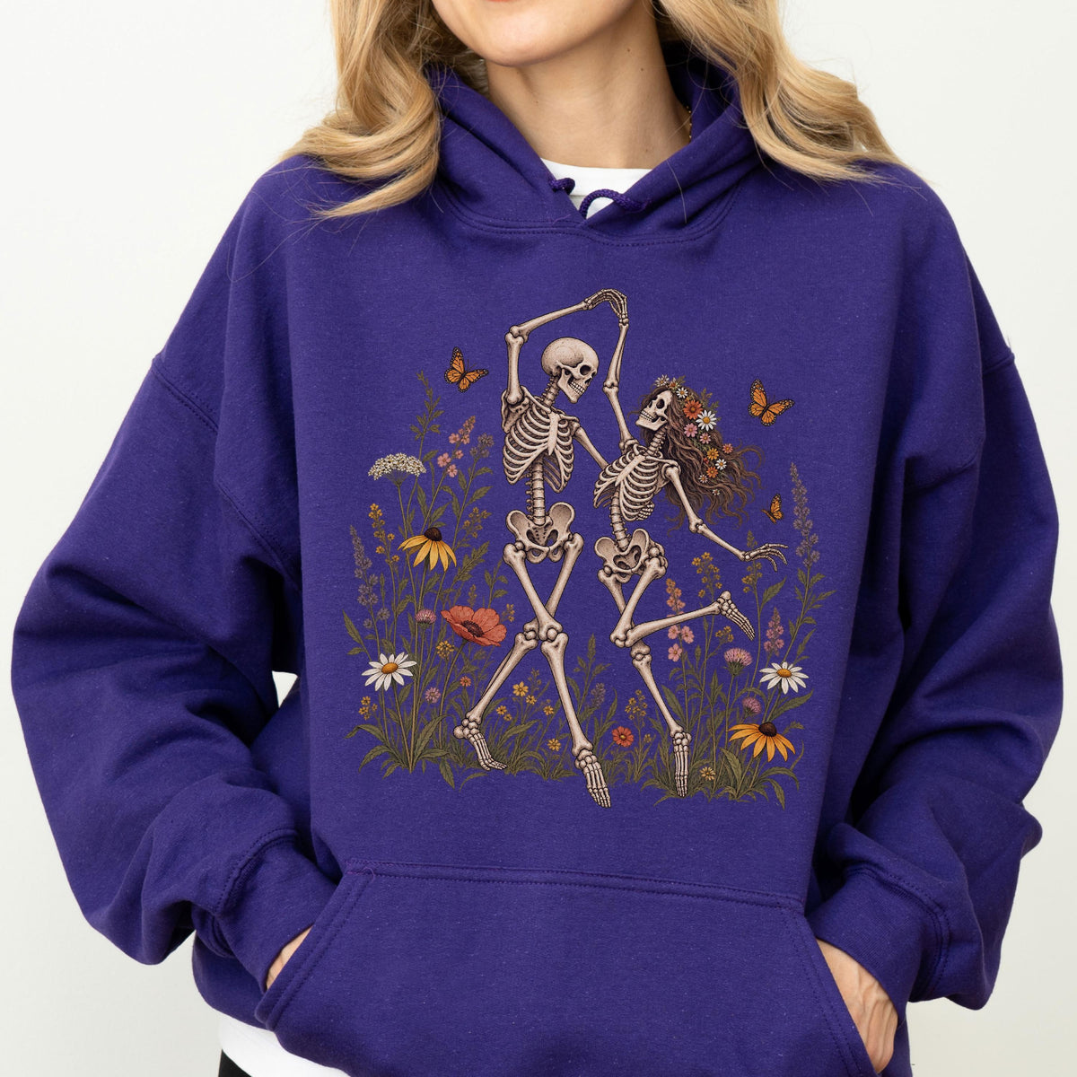 May include: A deep purple hooded sweatshirt featuring a graphic print of two skeletons dancing amidst a field of colorful wildflowers and orange butterflies. The hoodie includes long sleeves, a front kangaroo pocket, and a drawstring hood.