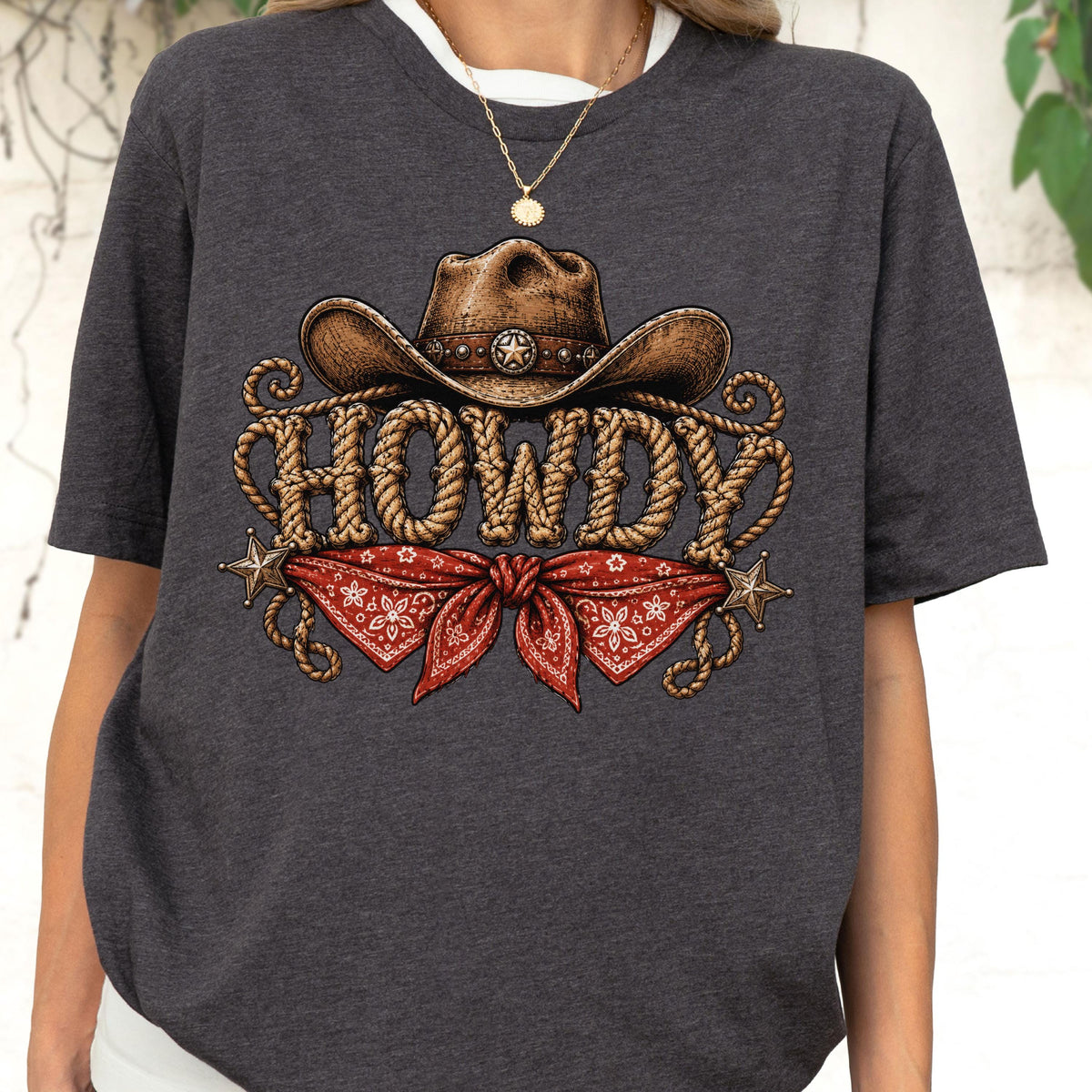 May include: A dark charcoal gray short-sleeve t-shirt featuring a graphic print of a brown cowboy hat above the word HOWDY in rope-style lettering, with a red patterned bandana tied below and two small metallic stars on the sides.