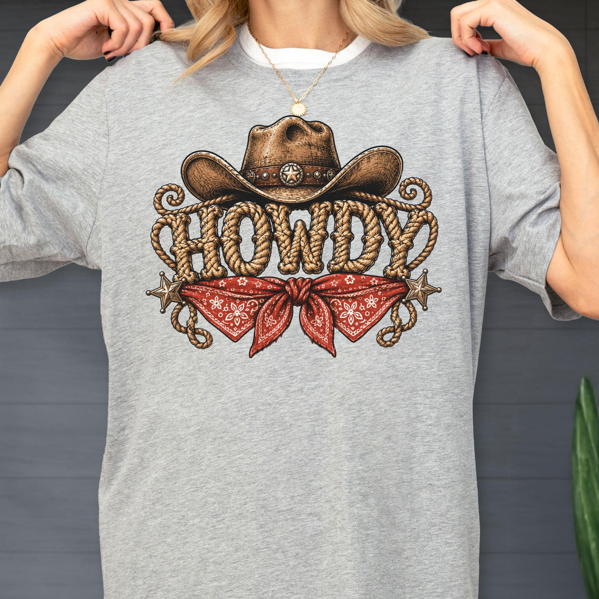 May include: A heather gray t-shirt featuring a graphic print of a brown cowboy hat, a red bandana with a white pattern, and the word HOWDY written in a rope-style font. Two small metallic-colored stars are positioned on either side of the bandana.