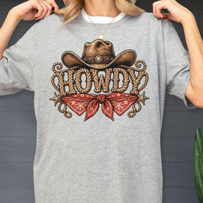 May include: A heather gray t-shirt featuring a graphic print of a brown cowboy hat, a red bandana with a white pattern, and the word HOWDY written in a rope-style font. Two small metallic-colored stars are positioned on either side of the bandana.