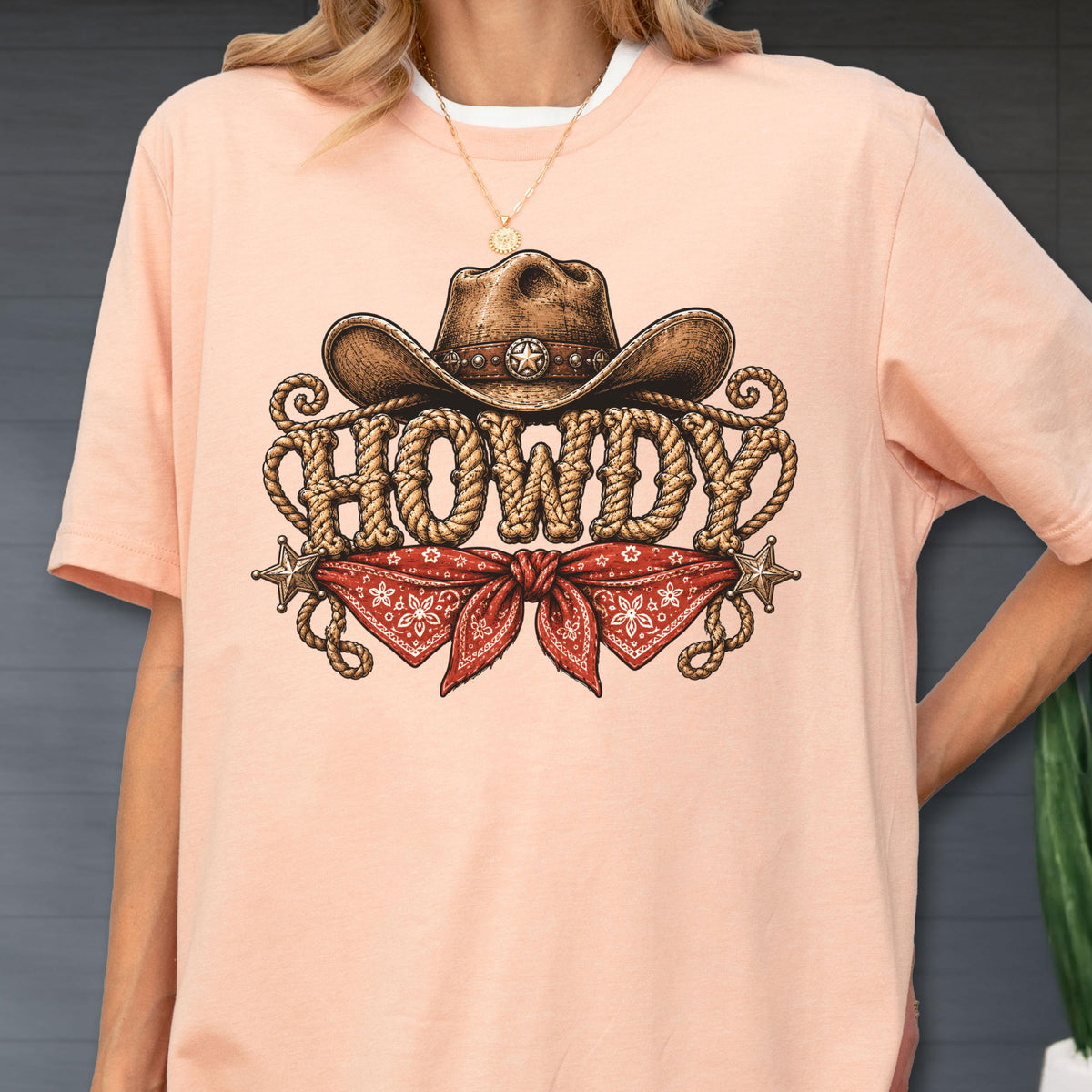 May include: A peach-colored t-shirt featuring a graphic print of a brown cowboy hat resting above the word HOWDY, which is written in a rope-style font. Below the text is a red bandana with a white pattern, flanked by two small silver stars.