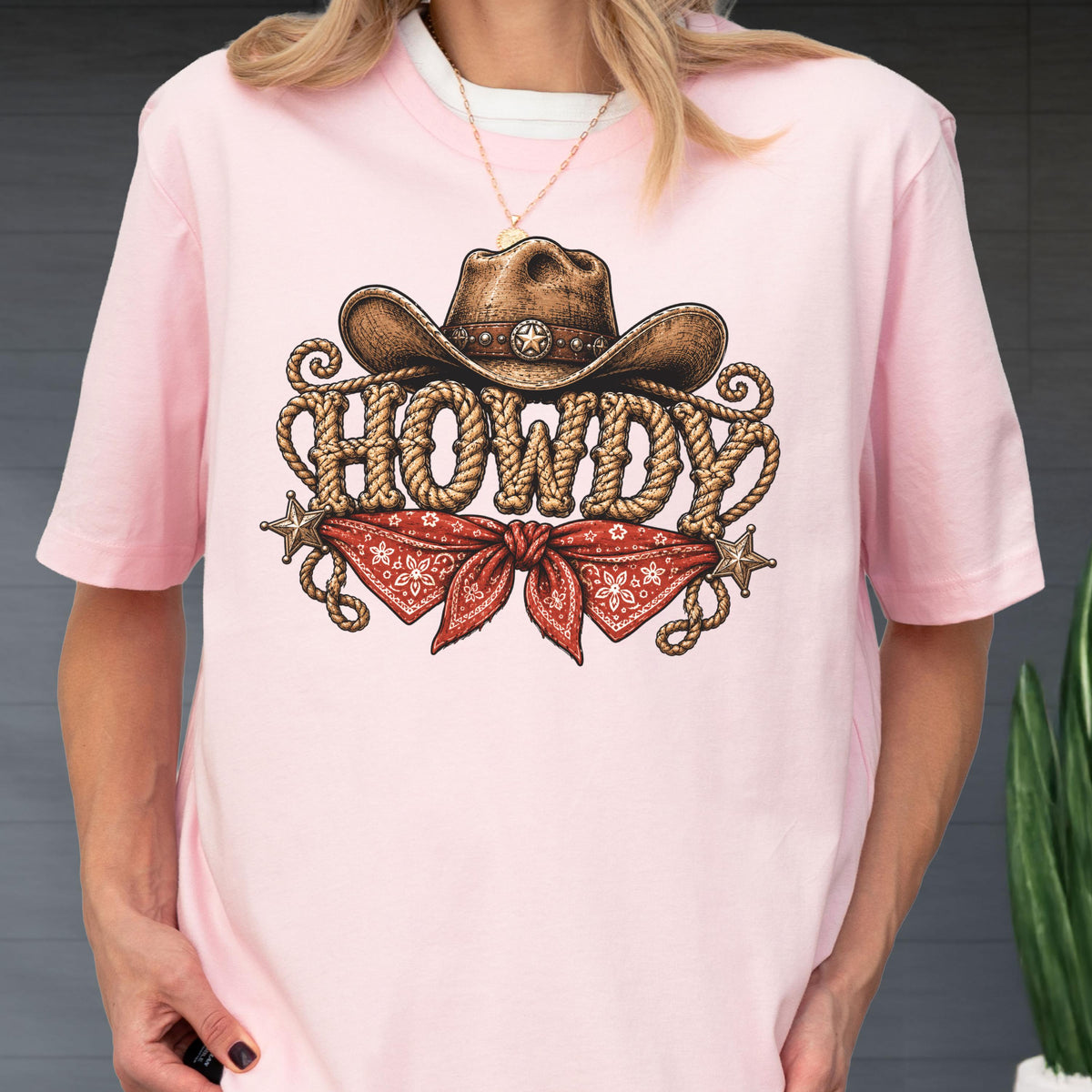 May include: A light pink t-shirt featuring a graphic print of a brown cowboy hat above the word HOWDY written in a rope-style font. Below the text is a red bandana with a white floral pattern, flanked by two small metallic star icons.