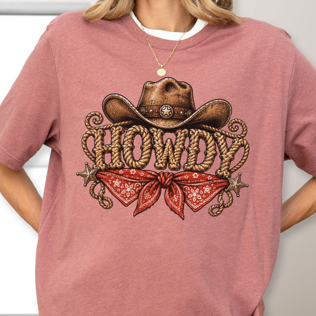 May include: A dusty rose t-shirt featuring a graphic print of a brown cowboy hat resting above the word HOWDY, which is written in a rope-style font. Below the text is a red bandana with a white floral pattern and two metallic star accents.