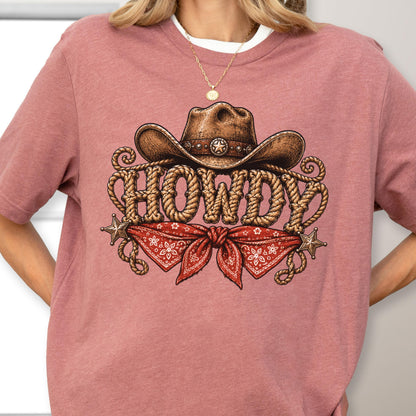 May include: A dusty rose t-shirt featuring a graphic print of a brown cowboy hat resting above the word HOWDY, which is written in a rope-style font. Below the text is a red bandana with a white floral pattern and two metallic star accents.
