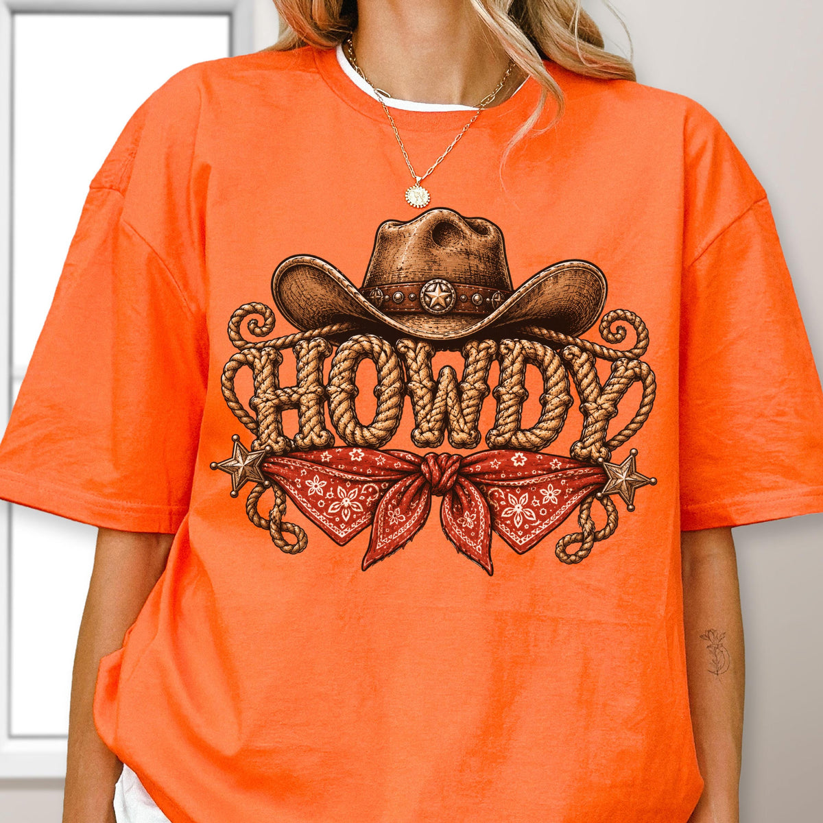 May include: A bright orange short-sleeve t-shirt featuring a graphic print of a brown cowboy hat, a red bandana with a white floral pattern, and the word HOWDY written in a rope-style font, accented by two small metallic-colored stars.