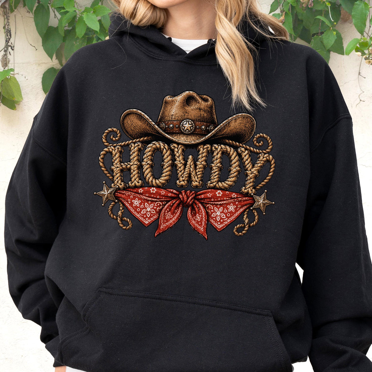May include: A black hooded sweatshirt featuring a graphic print of a brown cowboy hat above the word HOWDY written in a rope-like font. Below the text is a red bandana with a white floral pattern, flanked by two small metallic-colored stars.