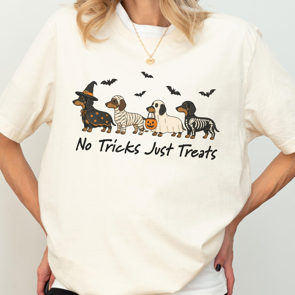May include: A cream-colored t-shirt featuring a graphic of four dachshunds in Halloween costumes, including a witch, a mummy, a ghost holding a pumpkin bucket, and a skeleton. The text "No Tricks Just Treats" is printed below the dogs.