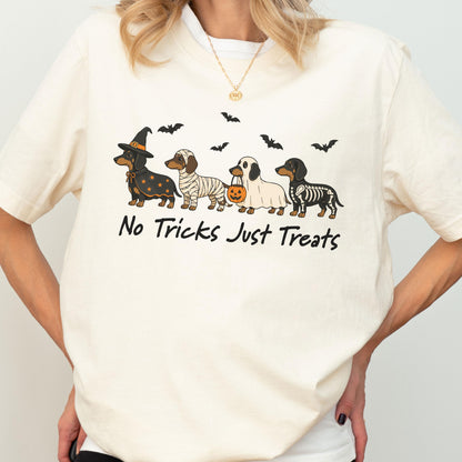May include: A cream-colored t-shirt featuring a graphic of four dachshunds in Halloween costumes, including a witch, a mummy, a ghost holding a pumpkin bucket, and a skeleton. The text "No Tricks Just Treats" is printed below the dogs.