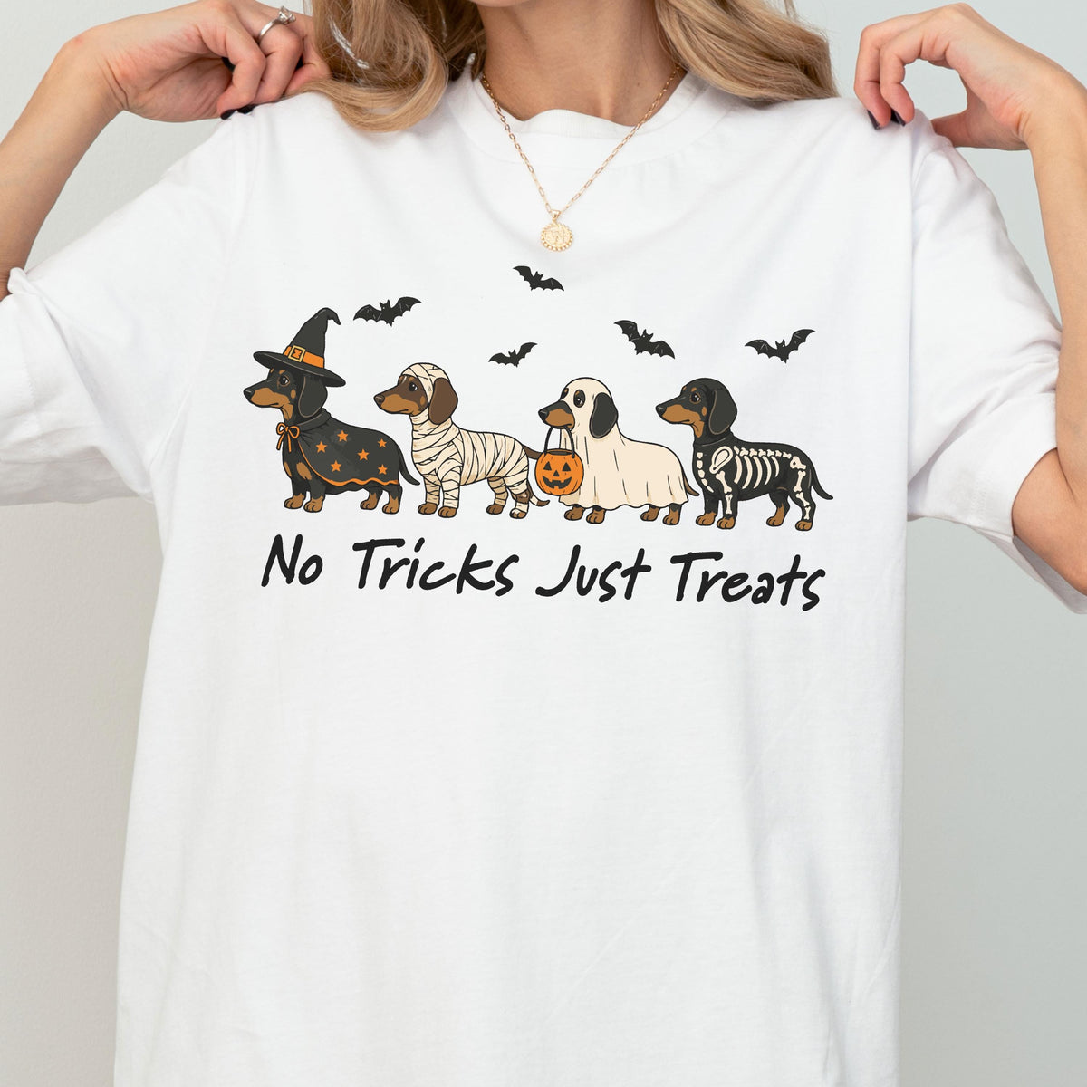 May include: A white t-shirt featuring a graphic of four dachshunds dressed in Halloween costumes: a witch, a mummy, a ghost holding a pumpkin bucket, and a skeleton. Above the dogs are black bats, and below is the text No Tricks Just Treats.