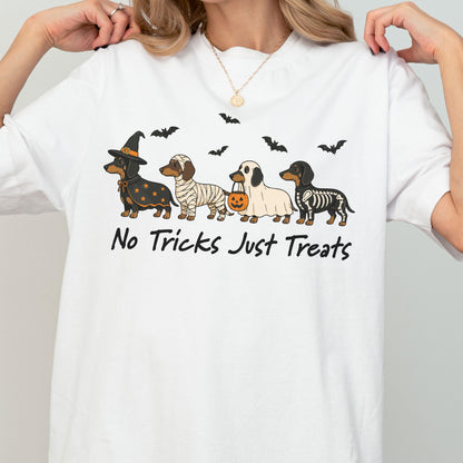 May include: A white t-shirt featuring a graphic of four dachshunds dressed in Halloween costumes: a witch, a mummy, a ghost holding a pumpkin bucket, and a skeleton. Above the dogs are black bats, and below is the text No Tricks Just Treats.