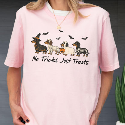 May include: A light pink short-sleeved t-shirt featuring a graphic of four dachshunds in Halloween costumes, including a witch, mummy, ghost, and skeleton, with bats and the text "No Tricks Just Treats" printed in black ink on the front.