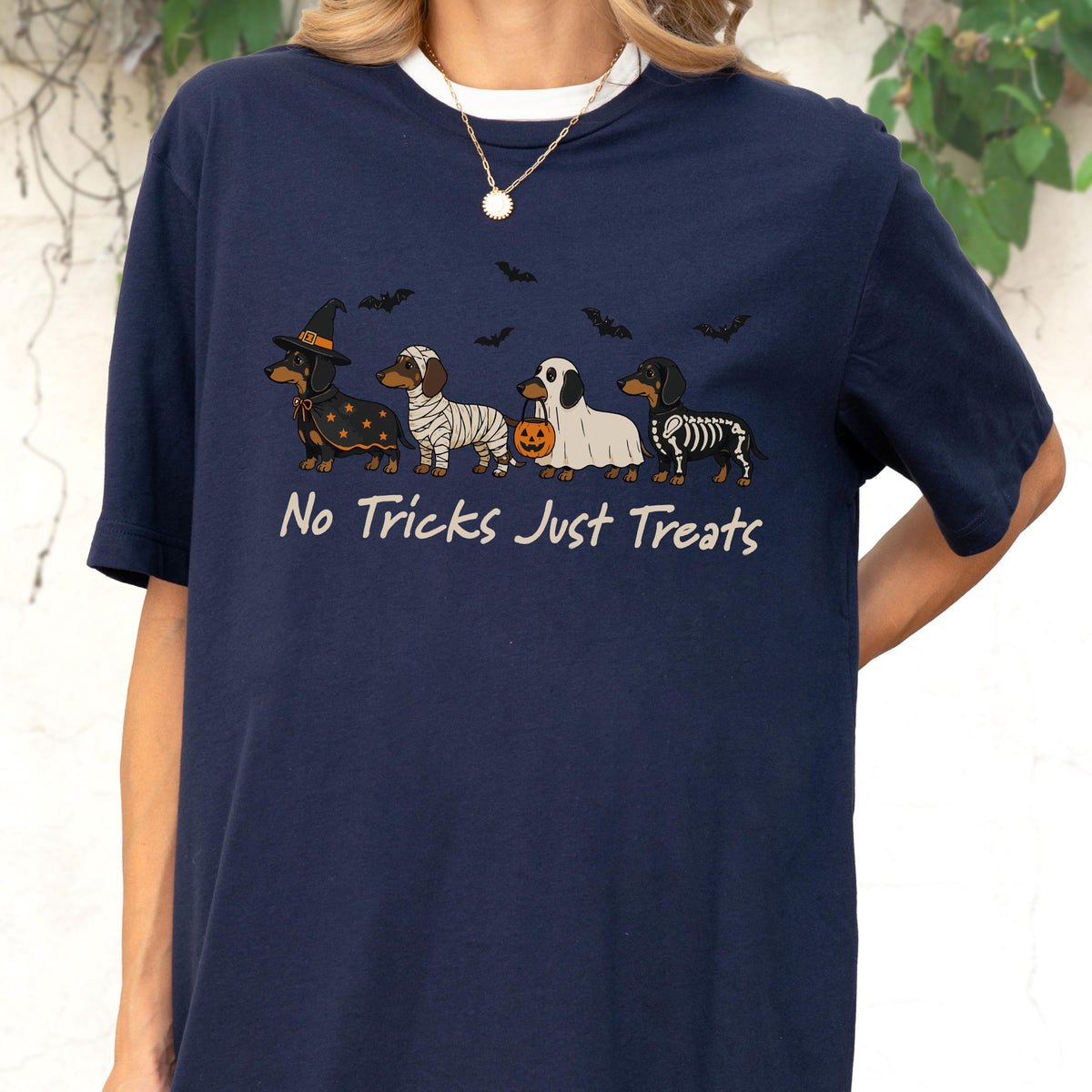 May include: A navy blue short-sleeve t-shirt featuring a graphic of four dachshunds in Halloween costumes, including a witch, mummy, ghost with a pumpkin bucket, and skeleton. The text "No Tricks Just Treats" is printed below the dogs.