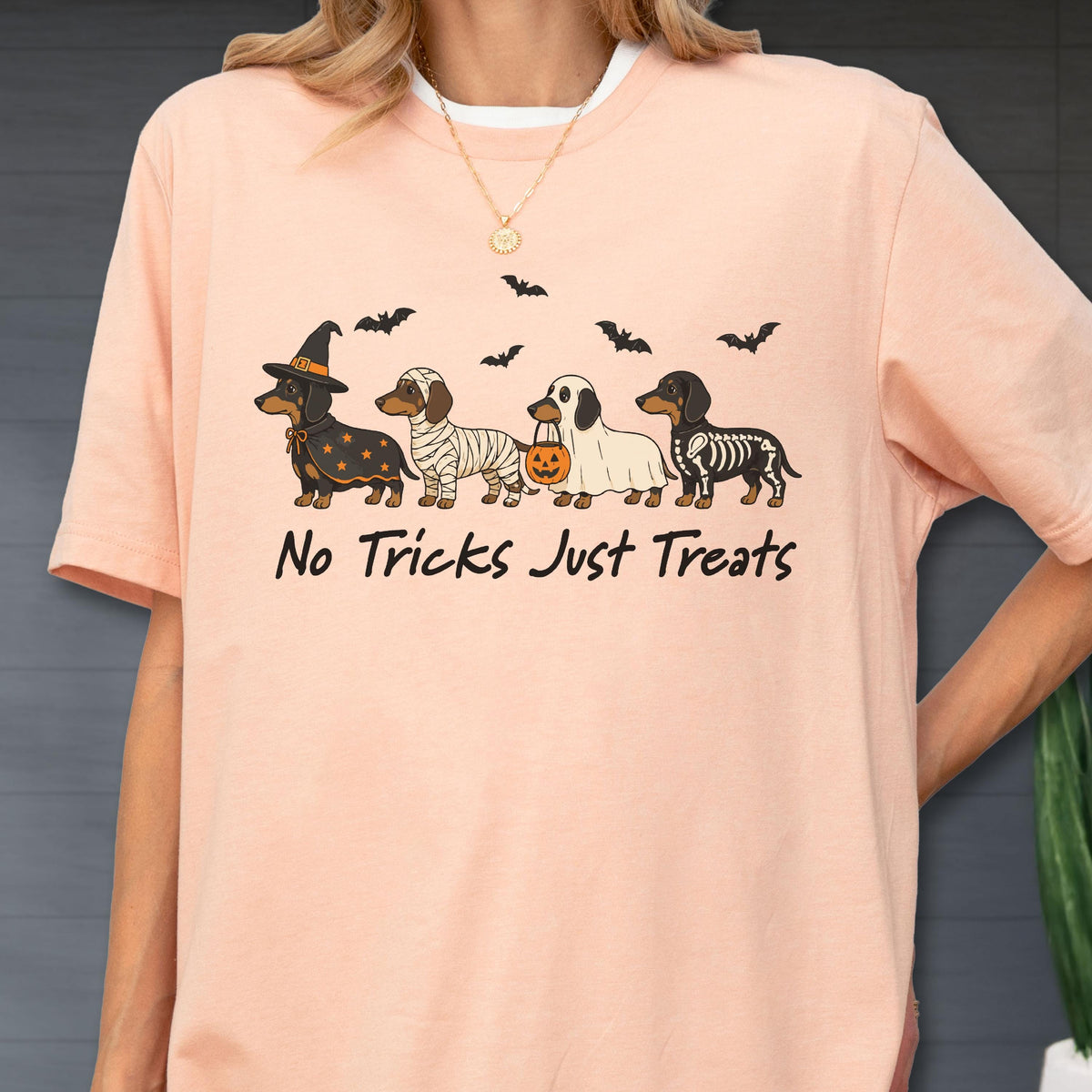 May include: A peach-colored t-shirt featuring a graphic of four dachshunds in Halloween costumes, including a witch, a mummy, a ghost with a pumpkin bucket, and a skeleton. Above the dogs are black bats, and the text reads No Tricks Just Treats.