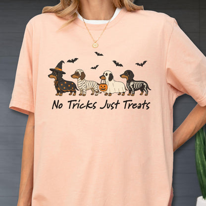 May include: A peach-colored t-shirt featuring a graphic of four dachshunds in Halloween costumes, including a witch, a mummy, a ghost with a pumpkin bucket, and a skeleton. Above the dogs are black bats, and the text reads No Tricks Just Treats.
