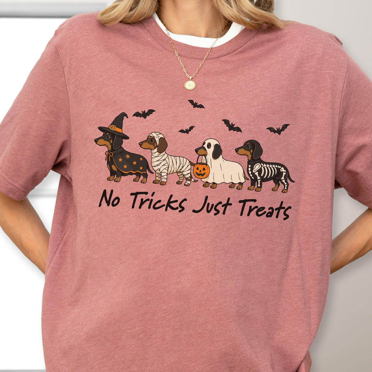 May include: A mauve t-shirt featuring a graphic of four dachshunds in Halloween costumes: a witch, a mummy, a ghost holding a pumpkin bucket, and a skeleton. Above the dogs are black bats. The text "No Tricks Just Treats" is printed below.