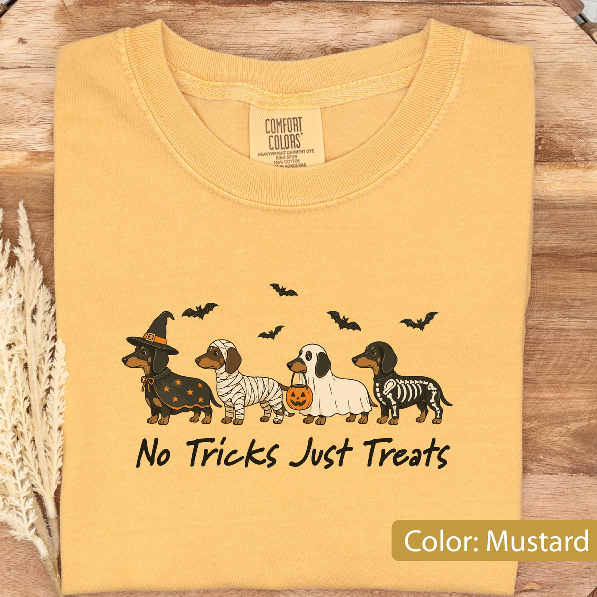 May include: A mustard yellow t-shirt featuring a graphic of four dachshunds in Halloween costumes: a witch, a mummy, a ghost holding a pumpkin bucket, and a skeleton. Above the dogs are black bats, and below is the text No Tricks Just Treats.