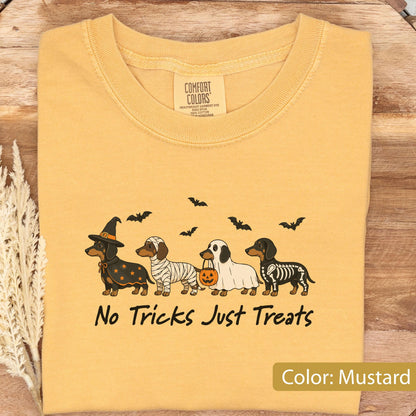 May include: A mustard yellow t-shirt featuring a graphic of four dachshunds in Halloween costumes: a witch, a mummy, a ghost holding a pumpkin bucket, and a skeleton. Above the dogs are black bats, and below is the text No Tricks Just Treats.