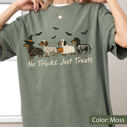 May include: A moss green t-shirt featuring a graphic of four dachshunds in Halloween costumes, including a witch, a mummy, a ghost holding a pumpkin bucket, and a skeleton. Above the dogs are black bats, and below is the text No Tricks Just Treats.