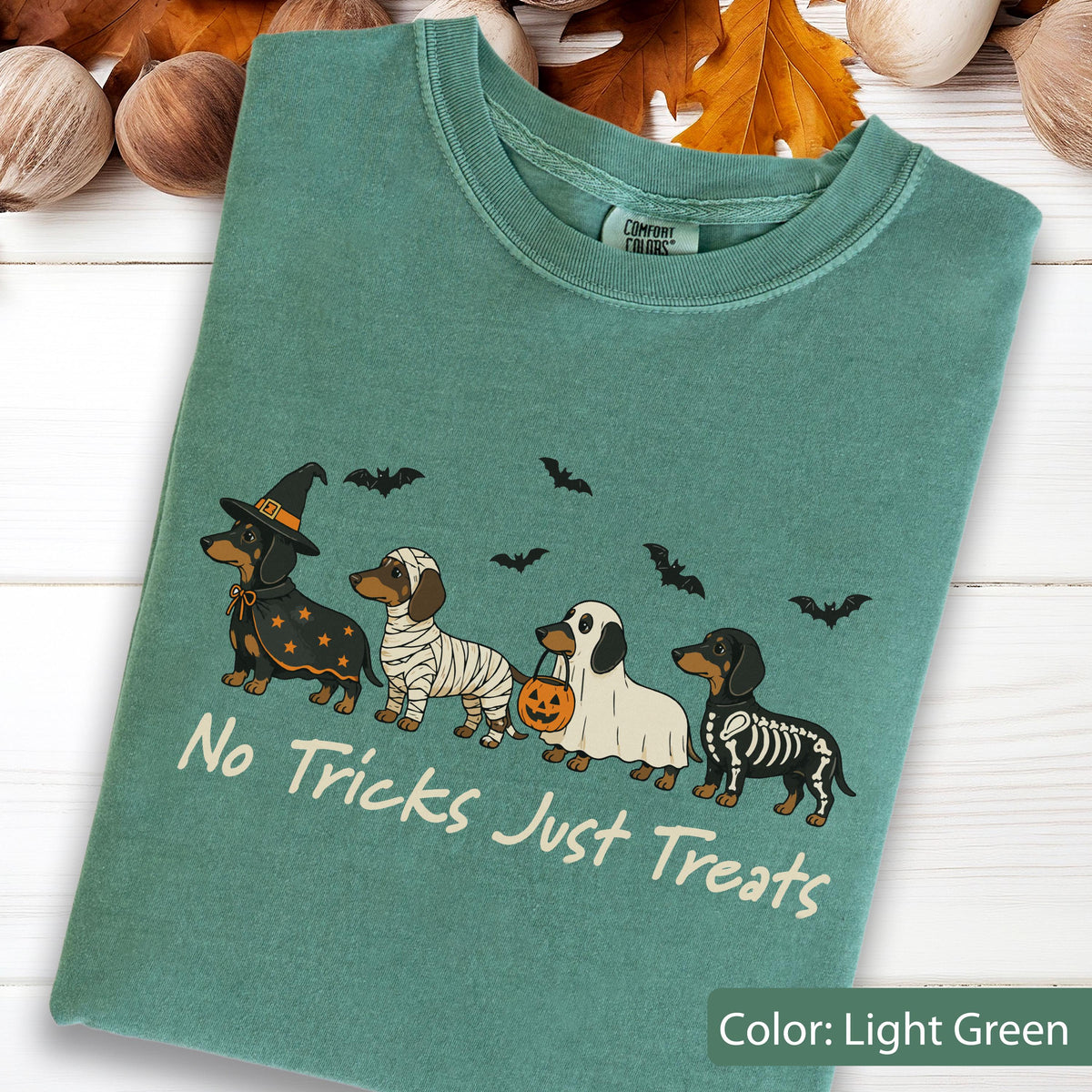 May include: A light green t-shirt featuring a graphic of four dachshunds in Halloween costumes, including a witch, a mummy, a ghost with a pumpkin bucket, and a skeleton. The text "No Tricks Just Treats" is printed below the dogs with bats above.