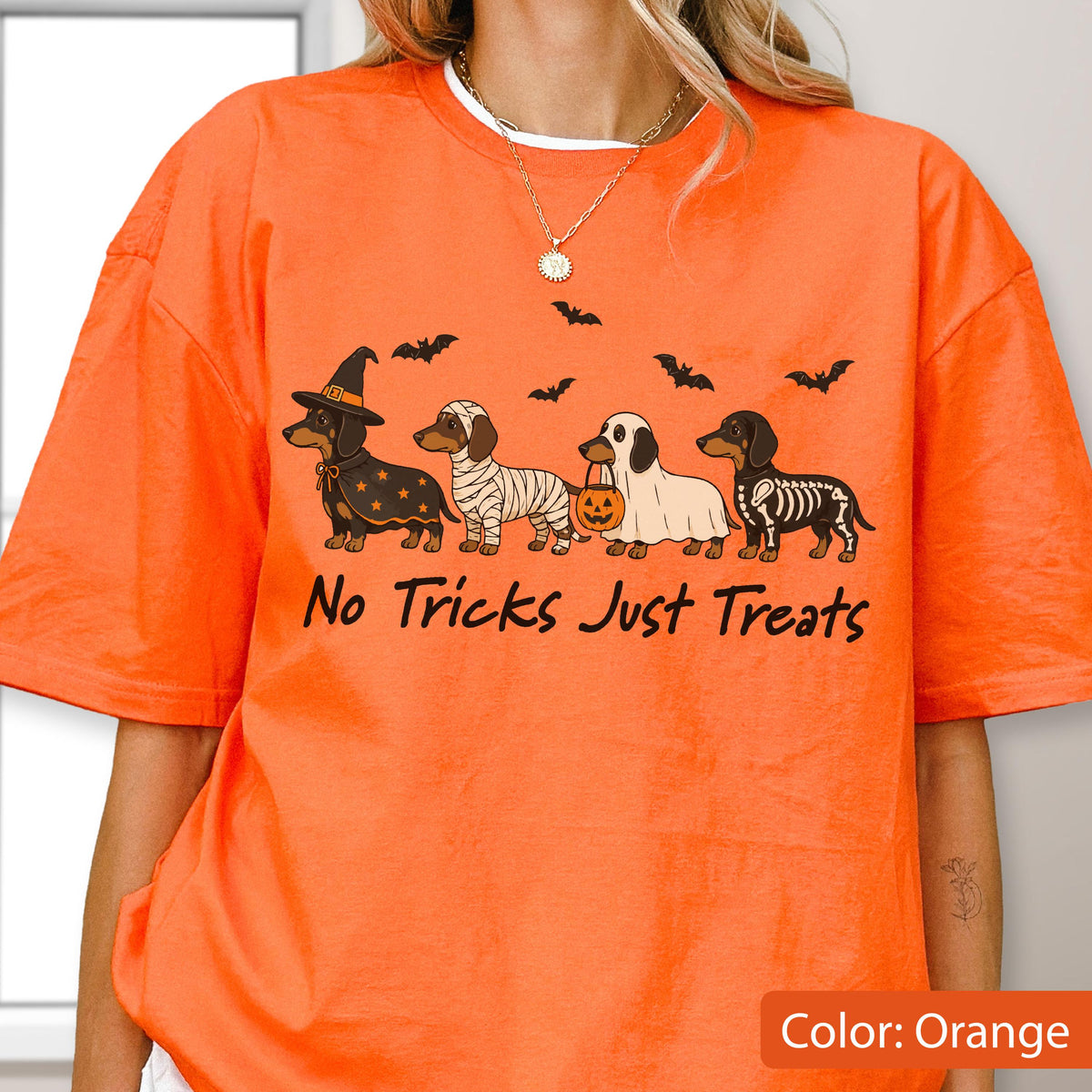 May include: An orange t-shirt featuring a graphic of four dachshunds in Halloween costumes, including a witch, a mummy, a ghost holding a pumpkin bucket, and a skeleton. The text "No Tricks Just Treats" is printed below the dogs with bats above.